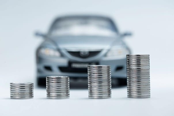 Imagem com um carro ao fundo e moedas emplhadas em primeiro plano